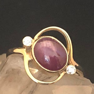 以下が含まれることがあります： 2 つの小さな透明な宝石で囲まれた、大きな楕円形の紫色の宝石が付いた金の指輪です。