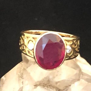 May include: A gold ring with a large oval ruby gemstone and two smaller diamond accents. The ring band is intricately designed with a filigree pattern.