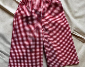 May include: A pair of trousers featuring a red and white gingham check pattern. The garment has a straight-leg silhouette and an elasticated waistband, providing a gathered texture at the top. The fabric is lightweight with a consistent grid.