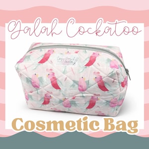 May include: A quilted cosmetic bag with a pink and white pattern featuring pink cockatoos and green leaves. The bag has a gray zipper and the text "Cosmetic Bag" in a tan font.