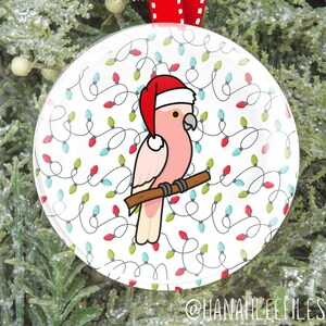 May include: A round Christmas ornament featuring a pink parrot wearing a Santa hat, perched on a brown branch. The background is white with a pattern of Christmas lights in red, green, and blue. A red ribbon is attached for hanging. The text "@HANAHLEEFILES" is at the bottom.