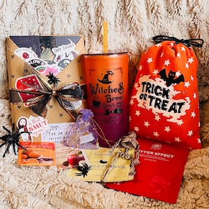May include: A Halloween-themed gift set featuring an orange and purple tumbler with "Witches Brew" text, a "Trick or Treat" bag, a wrapped package with stickers, and various small items like eye masks and tea.