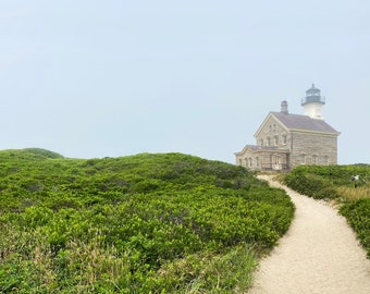 May include: A stone lighthouse with a white tower stands on a grassy hill with a path leading up to it. The sky is overcast and foggy.
