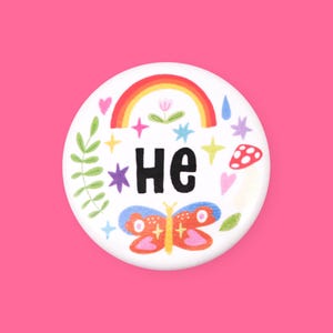 Pode incluir: Botão branco com um arco-íris, estrelas, uma borboleta e a palavra "He" em letras pretas.
