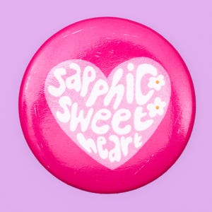 May include: A round, hot pink button with the words "Sapphic Sweetheart" in white script inside a white heart. Small white flowers with yellow centers are on the right side of the heart. The button has a glossy finish.