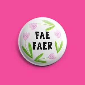 May include: White button with the words "FAE FAER" in black, surrounded by pink and green floral illustrations. The button is set against a bright pink background.