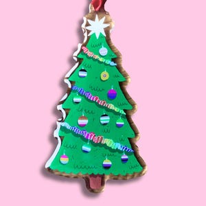 Op de afbeelding: Een groene kerstboomversiering met een ster en een rood lint om op te hangen. De boom is versierd met kleurrijke ballen en een slinger. De versiering is gemaakt van hout en heeft een witte omtrek. De boom is ongeveer 10 cm hoog.