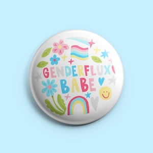 Könnte beinhalten: Ein weißer Button mit den Worten "GENDERFLUX BABE" in bunten Buchstaben. Der Button zeigt einen Regenbogen, Blumen, Sterne und ein lächelndes Gesicht. Das Design ist vor einem hellblauen Hintergrund platziert.
