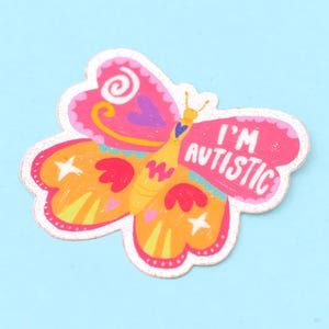 Könnte beinhalten: Ein rosa und gelber Schmetterlingsaufkleber mit dem Text "I'm Autistic" auf dem rechten Flügel. Der Schmetterling hat einen weißen Umriss und Glitzerakzente.