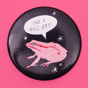Puede incluir: Un bot&oacute;n negro con una rana rosa y una burbuja de di&aacute;logo blanca que dice "Have a nice day!" El fondo es negro con estrellas blancas.