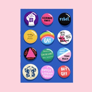 Op de afbeelding: Een verzameling kleurrijke ronde buttons met diverse teksten en afbeeldingen. De buttons tonen zinnen als "GAY", "TRANS PRIDE" en "SUPPORT LESBIAN FAMILIES". De achtergrond is effen blauw, met een roze rand.