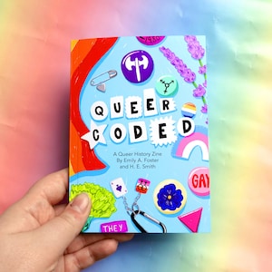 Puede incluir: Un zine azul y rojo titulado "Queer Coded: A Queer History Zine" de Emily A. Foster y H. E. Smith. La portada presenta un arcoíris, una flor de lavanda, un alfiler de seguridad, un martillo y una hoz, y una variedad de otros pines coloridos.