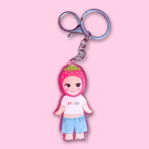May include: A keychain with a cartoon character in a strawberry hat and blue shorts. The character wears a white t-shirt with the word "fruity" in rainbow colours. The keychain has a silver clasp and ring.