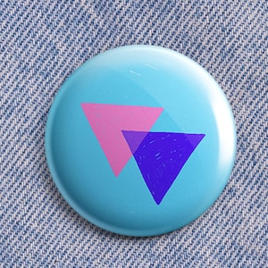 May include: A round, light blue button with a glossy finish. The button features two overlapping triangles, one pink and one purple. The button is set against a denim background.