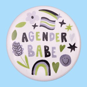 May include: White button featuring the text "Agender Babe" in black, green, and grey lettering. The button is adorned with a rainbow, stars, hearts, and flowers.