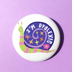 Puede incluir: Un botón blanco con una ilustración caprichosa de un caracol. El caracol tiene un cuerpo verde con rayas rosas y verdes, y una concha azul con las palabras "I'M DYSLEXIC" en blanco. La concha está decorada con estrellas y una flor.