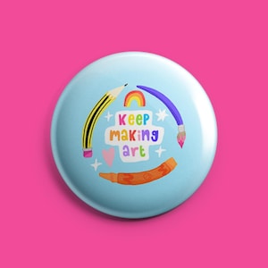 May include: A light blue badge with the text "Keep Making Art" and illustrations of art supplies. The design includes a pencil, paintbrush, rainbow, heart, and crayon in various colours. The badge is set against a bright pink background.