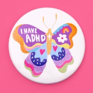 Peut inclure: Un bouton blanc avec un motif de papillon coloré. Le papillon a des ailes violettes avec le texte "I HAVE ADHD" sur une aile. Le papillon a également un arc-en-ciel, une fleur et des cœurs sur ses ailes.