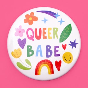 Może przedstawiać: Biały guzik z tęczą, sercami, gwiazdami, kwiatami i liśćmi. Tekst "QUEER BABE" jest napisany w kolorach tęczy.