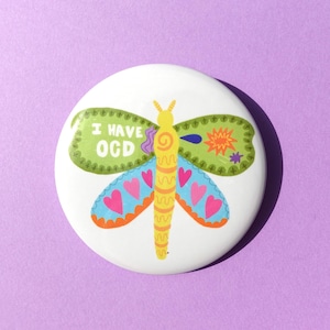 Puede incluir: Un botón blanco con un diseño de libélula colorida. La libélula tiene alas verdes con el texto "I HAVE OGD", alas azules y naranjas con corazones rosas y un cuerpo amarillo. Sobre un fondo morado.