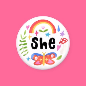 Puede incluir: Botón blanco con un arcoíris, una mariposa, estrellas, corazones, hojas y setas. El texto "She" está en letras negras.