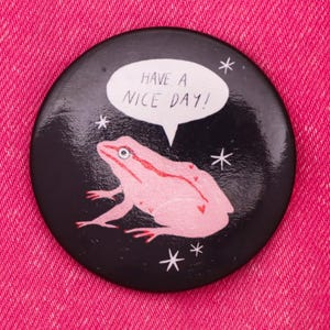 Puede incluir: Un bot&oacute;n negro con una rana rosa y una burbuja de di&aacute;logo blanca que dice "Have a nice day!" El fondo es negro con estrellas blancas.