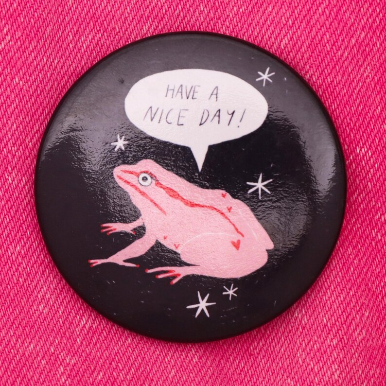 Puede incluir: Un bot&oacute;n negro con una rana rosa y una burbuja de di&aacute;logo blanca que dice "Have a nice day!" El fondo es negro con estrellas blancas.