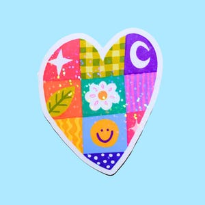 Puede incluir: Una pegatina en forma de corazón con un diseño de patchwork colorido. El diseño incluye una estrella, una hoja, una flor, una carita sonriente y una luna creciente. La pegatina tiene un borde blanco y un fondo azul claro.