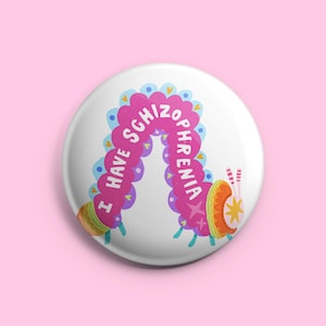 Puede incluir: Botón blanco con un diseño de oruga colorida. La oruga es rosa, azul y amarilla, con el texto "I HAVE SCHIZOPHRENIA" en blanco. Sobre un fondo rosa.