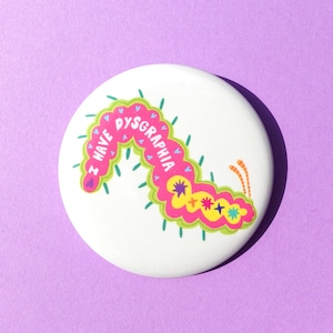 Puede incluir: Un botón blanco con una ilustración colorida de una oruga. La oruga es rosa, amarilla y verde, con las palabras "I HAVE DYSGRAPHIA" en rosa. El botón está sobre un fondo morado.