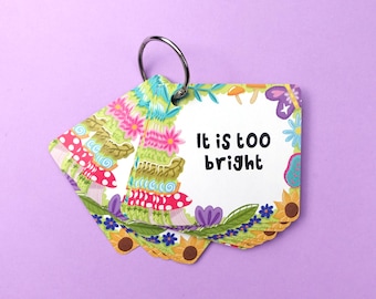 Puede incluir: Un conjunto de tarjetas ilustradas coloridas, sujetas por un anillo plateado. Las tarjetas presentan un diseño floral y de setas con el texto "It is too bright" sobre un fondo blanco. El fondo es de color púrpura liso.