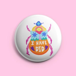 Op de afbeelding: Witte knoop met een kleurrijke illustratie van een gestileerde kever. De kever heeft een regenboogkop, stervormige ogen en de tekst "I HAVE DID" in oranje. De achtergrond is effen roze.