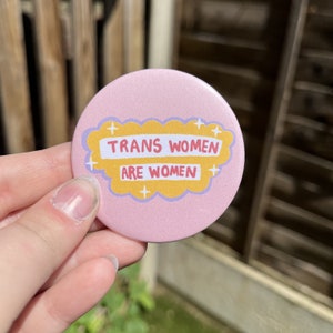 May include: Pink button with a yellow cloud and the text "Trans women are women" in white.