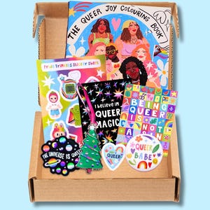 Könnte beinhalten: Ein Karton voller Artikel zum Thema Pride. Enthält ein Malbuch mit dem Titel "The Queer Joy Colouring Book", Aufkleber, einen Weihnachtsbaumschmuck, einen herzförmigen Aufkleber und eine Anstecknadel mit der Aufschrift "Queer Babe".