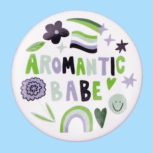 May include: A white button with the words "AROMANTIC BABE" in green and purple lettering. The button features various illustrations, including a flower, a rainbow, stars, and hearts, all in shades of green, purple, and black.