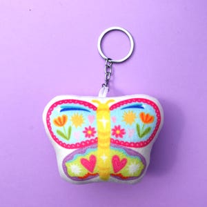 Puede incluir: Un llavero blanco con forma de mariposa con un diseño floral colorido. La mariposa tiene un fondo azul claro con flores naranjas, soles amarillos y corazones rosas. El llavero tiene un anillo y una cadena plateados.