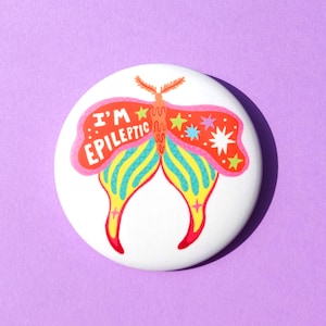 Puede incluir: Un botón redondo blanco con un diseño de mariposa colorido. La mariposa tiene alas rojas, naranjas, amarillas, azules y rosas, con el texto "I'M EPILEPTIC" en rojo. El botón está sobre un fondo morado.