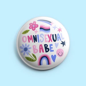 Peut inclure: Un badge blanc avec l'inscription "OMNISEXUAL BABE" en rose et bleu, orné d'illustrations de fleurs, d'étoiles, d'un arc-en-ciel et d'un smiley. Le badge est posé sur un fond bleu clair.