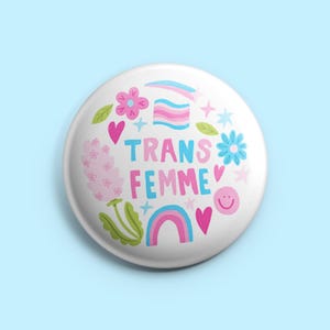 Peut inclure: Un badge blanc avec l'inscription "TRANS FEMME" en bleu et rose, entouré d'illustrations colorées de fleurs, de cœurs, d'un arc-en-ciel et d'un drapeau de la fierté transgenre. Le badge a une finition brillante.