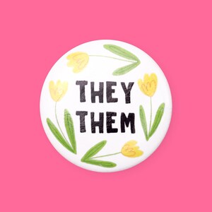 May include: White button with a black and white graphic that says "They Them" surrounded by yellow flowers with green stems on a white background.