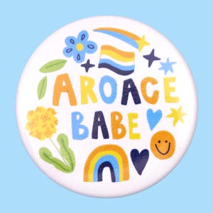 May include: A white button with a colorful design featuring the text "aroace babe" surrounded by rainbows, stars, hearts, and flowers.