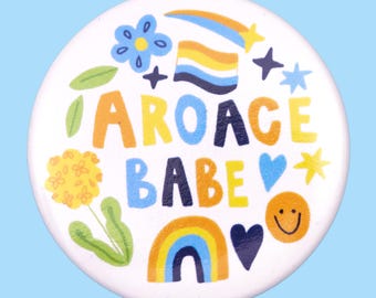 May include: A white button with a colorful design featuring the text "aroace babe" surrounded by rainbows, stars, hearts, and flowers.