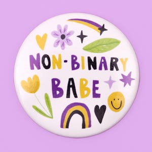 Puede incluir: Botón blanco con el texto "NON-BINARY BABE" en letras negras, moradas y amarillas. El botón está decorado con un arcoíris, estrellas, corazones, flores y una carita sonriente.