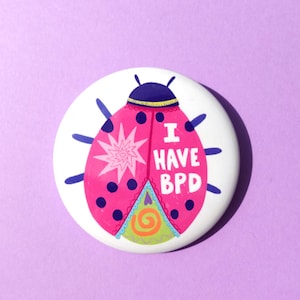 Op de afbeelding: Een ronde, witte button met een roze lieveheersbeestje ontwerp. Het lieveheersbeestje heeft zwarte stippen en blauwe antennes. De tekst "I HAVE BPD" staat in het wit. De achtergrond is lavendelkleurig.