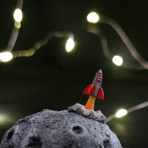 Puede incluir: Un pequeño cohete rojo y gris despega de una luna gris texturizada. Llamas naranjas emergen de la base. Luces de cadena amarillas suaves en el fondo.