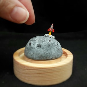 Puede incluir: Miniatura de luna gris con cráteres, un cohete rojo y plateado despegando y escape amarillo. La luna se asienta sobre una base de madera. La imagen es un primer plano.