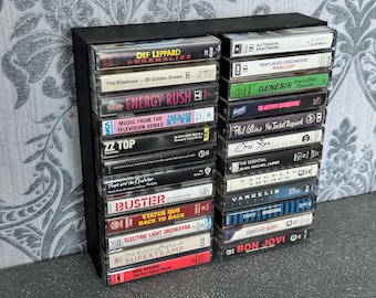 May include: A black cassette tape storage rack with 20 cassette tapes. The tapes are arranged vertically in two columns. The tapes have various artist names and album titles, including Def Leppard, The Shadows, Energy Rush, ZZ Top, Phil Collins, Genesis, and Bon Jovi.