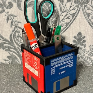 May include: A blue and red desk organiser made from recycled floppy disks. The organiser is filled with various office supplies, including scissors, pens, pencils, and a glue stick. The text on the floppy disk reads "DUNE II AMIGA UK/FRENCH GERMAN Disk 3 of 5".