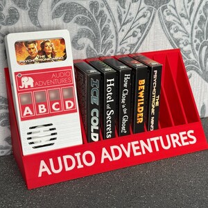May include: A red plastic display stand with the text "AUDIO ADVENTURES" holds a white audio player and several black cassette tapes. The audio player has the text "AUDIO ADVENTURES" and the letters A, B, C, and D.