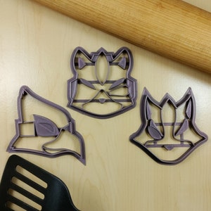 May include: Three purple plastic cookie cutters in the shape of video game character faces. A wooden rolling pin and a black spatula are also visible. The cookie cutters are on a light wood surface.